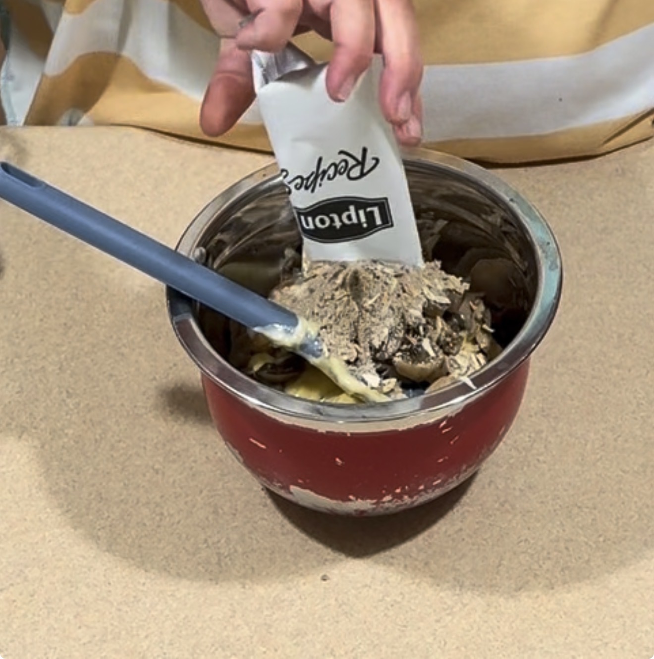 A person pours a packet of Lipton Recipe Secrets onion soup mix into a bowl with other ingredients, using a blue spatula to mix.
