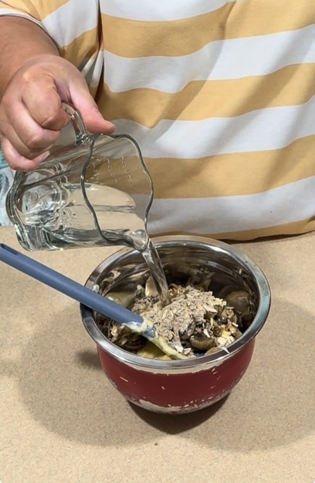 A person pours water from a glass pitcher into a metal mixing bowl filled with oats and chopped mushrooms, using a blue spatula to stir.