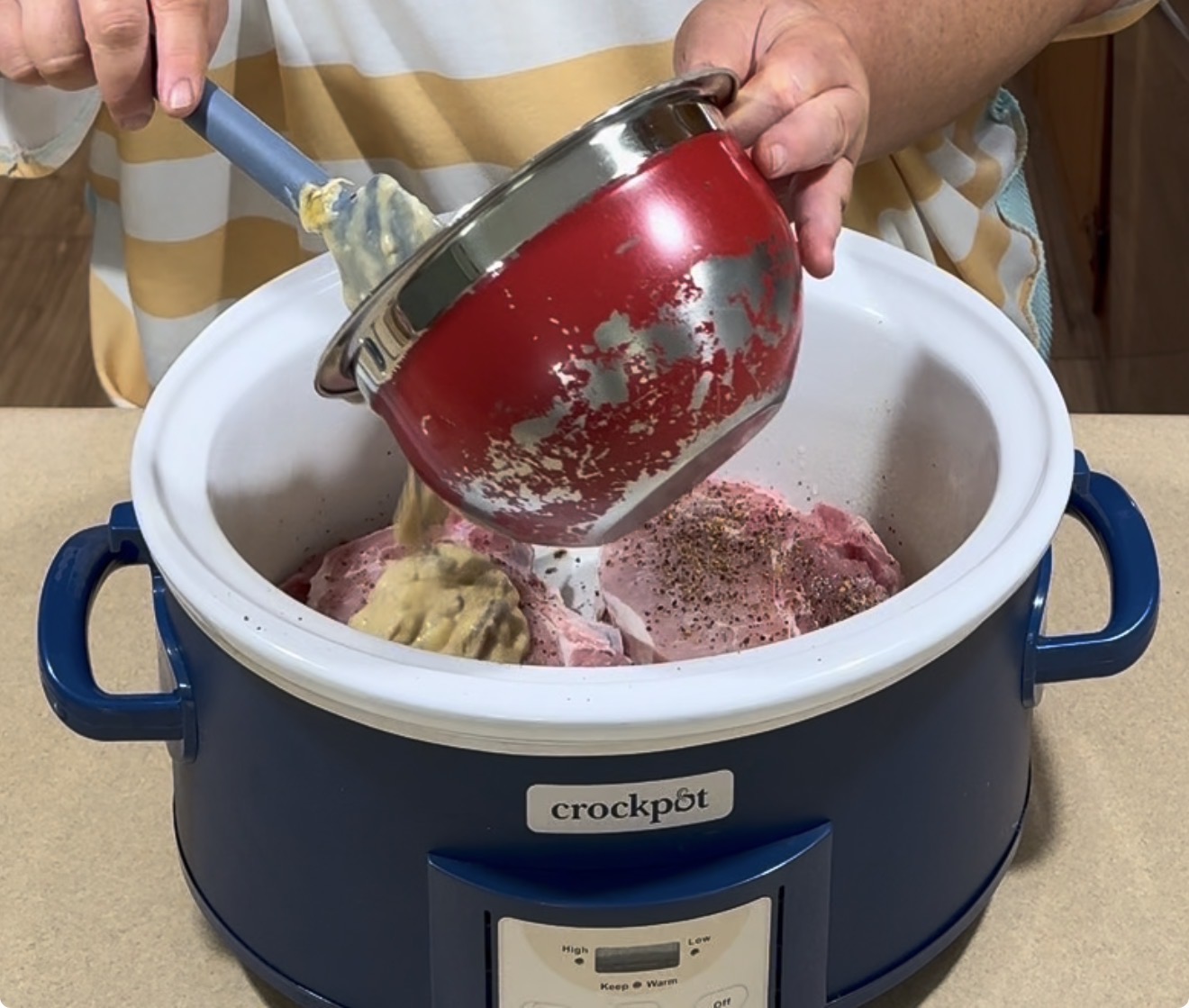 A person pours a mixture from a red saucepan into a blue Crockpot filled with meat and spices while standing at a kitchen counter.