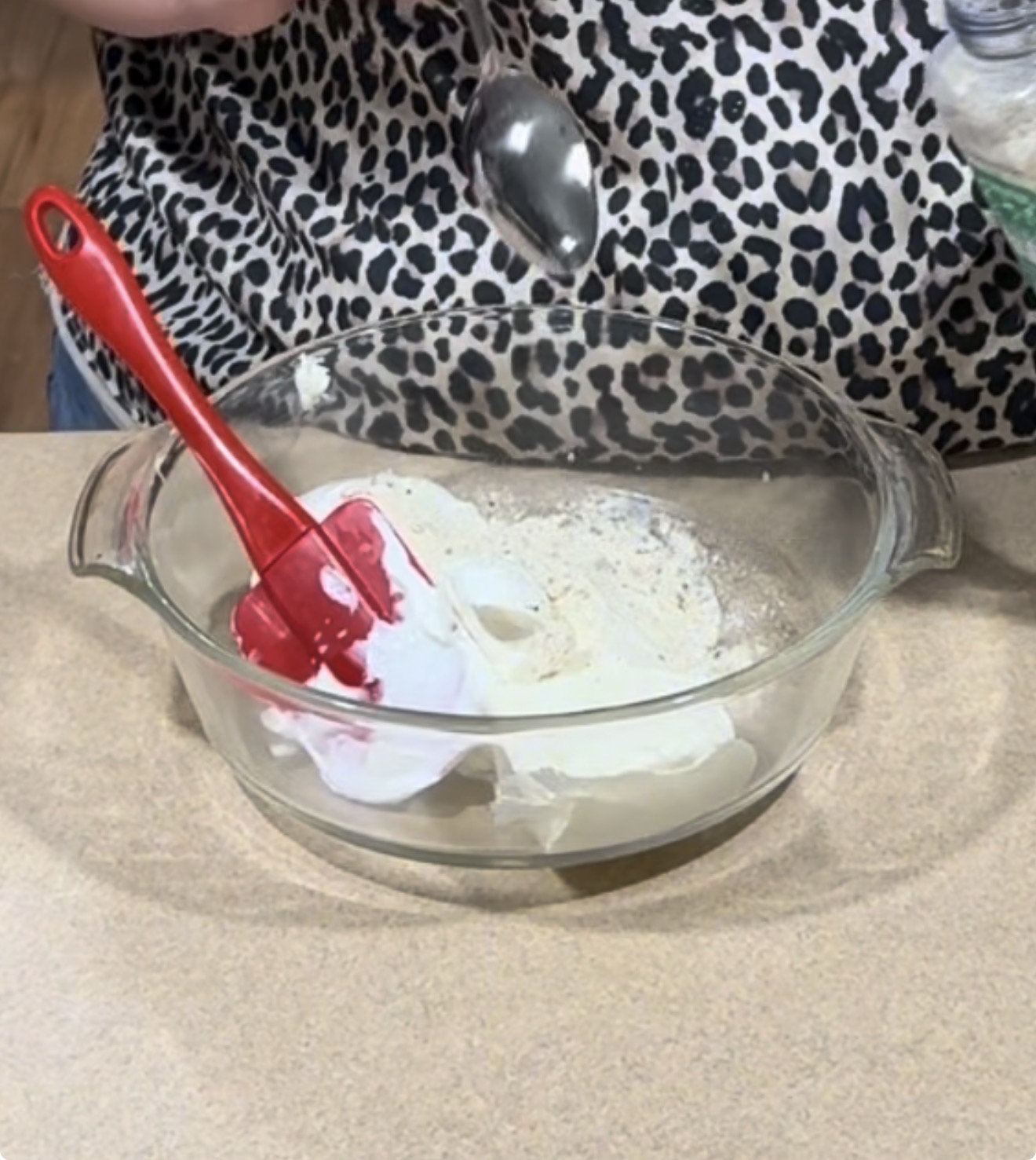 A person in a leopard print shirt is mixing white powder and other ingredients in a glass bowl with a red spatula, while holding a spoon above the bowl.