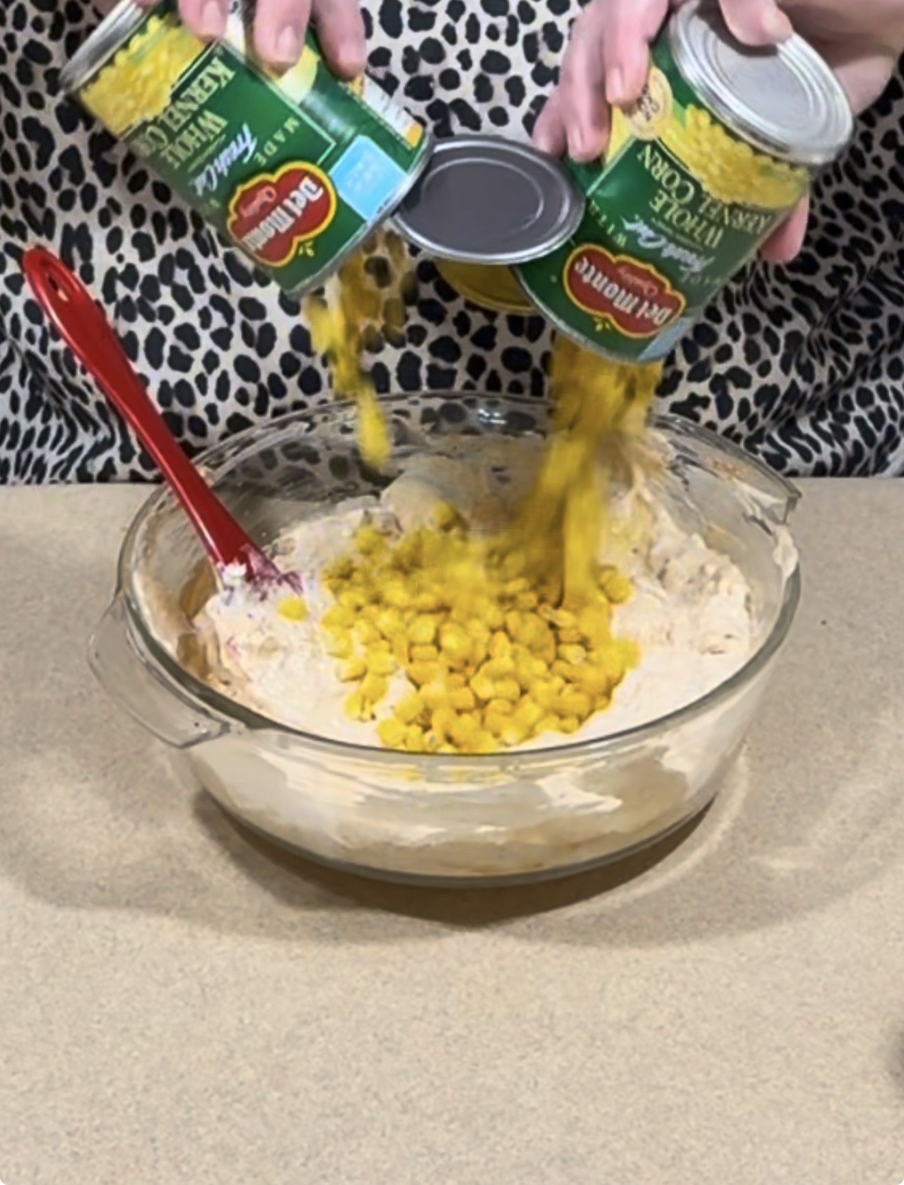 Two cans of drained corn are being poured into a glass mixing bowl filled with a creamy mixture. A red spatula rests in the bowl, and a person in a spotted shirt holds the cans over the bowl.