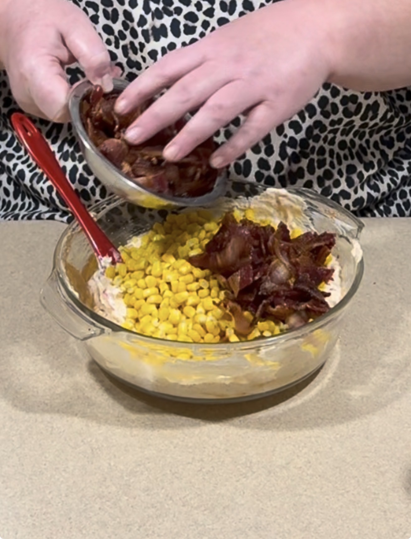 A person adds cooked bacon pieces to a glass bowl containing corn and a creamy mixture, with a red spatula beside the ingredients on a beige countertop.