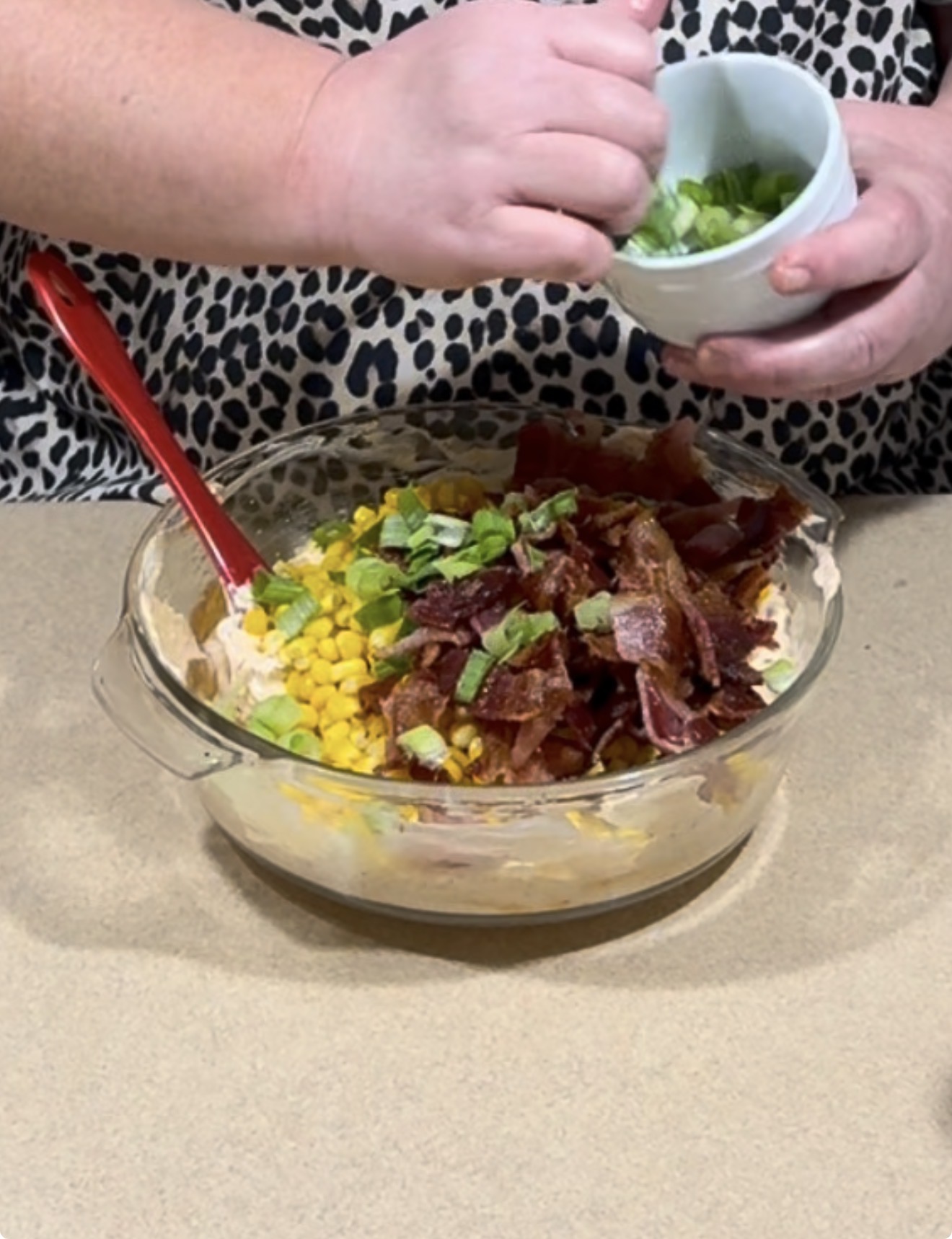 A person sprinkles chopped green onions from a small bowl onto a glass dish filled with corn, bacon, and other ingredients, preparing a mixed dish on a kitchen counter.
