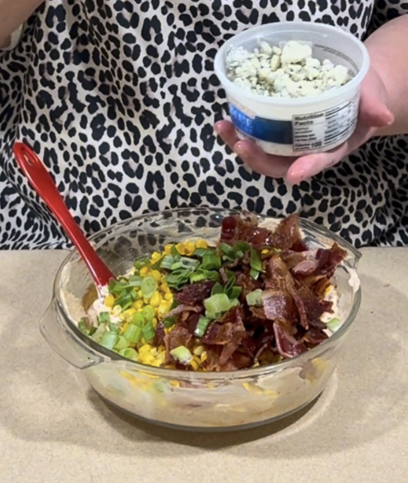 A person in a leopard print shirt holds a container of blue cheese above a mixing bowl with bacon, corn, green onions, and a red spoon.