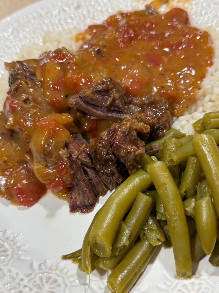A plate of pot roast with tomato-based gravy served over rice, alongside a portion of cooked green beans.