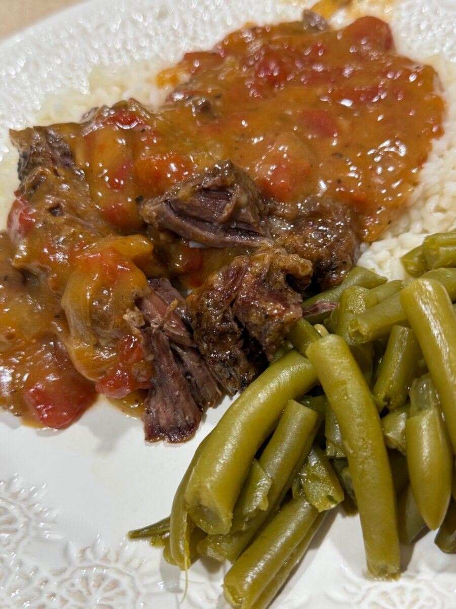 A plate of pot roast with tomato-based gravy served over rice, alongside a portion of cooked green beans.