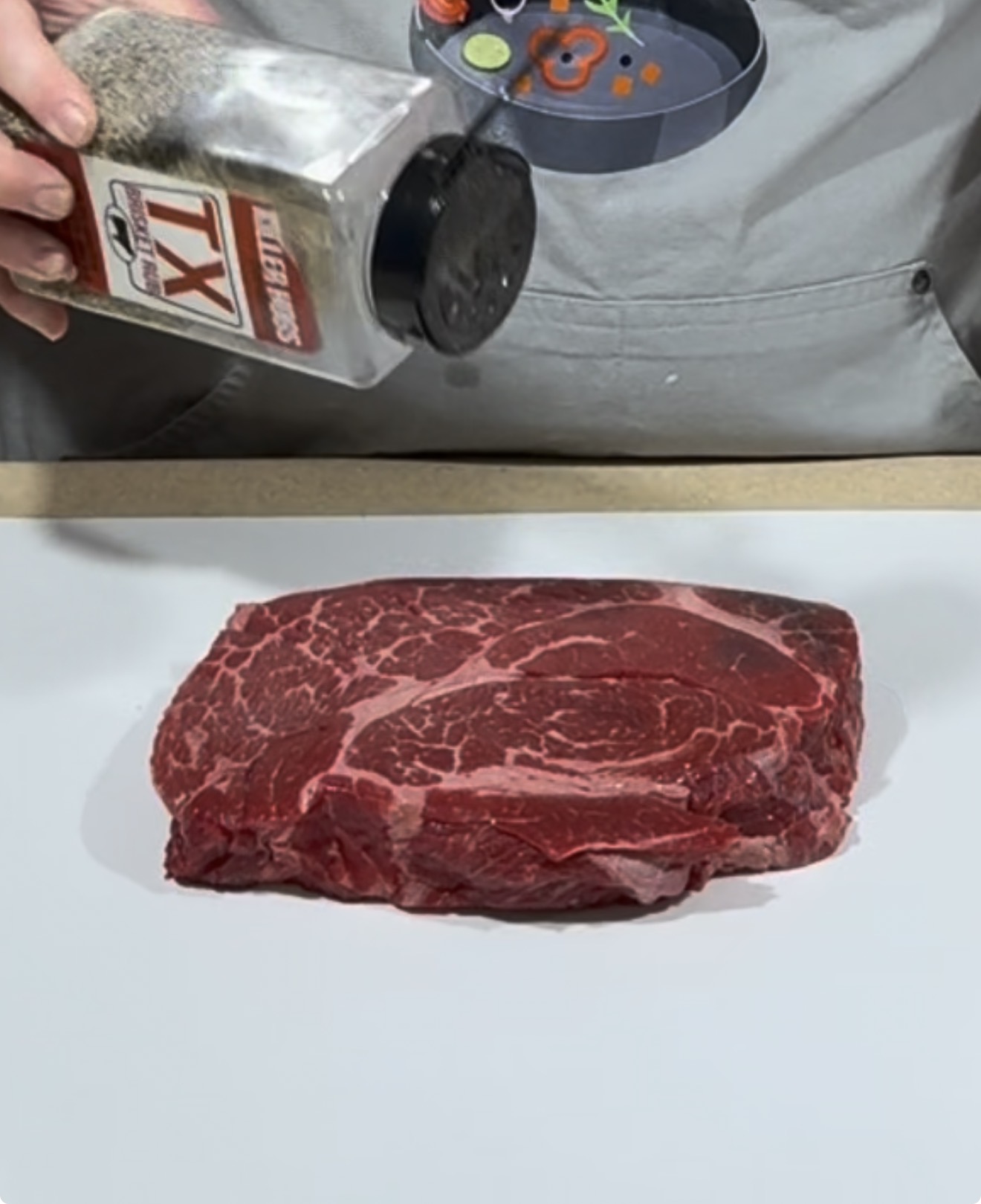 A person sprinkles TX seasoning from a container onto a raw marbled steak placed on a white surface. A gray apron with a colorful design is partially visible in the background.
