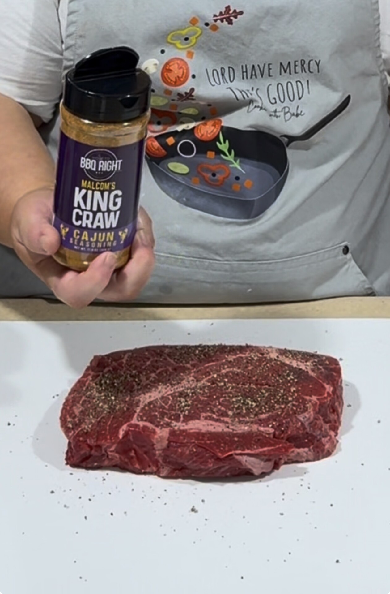 A person wearing a cooking-themed apron holds BBQ Right King Craw Cajun seasoning above a raw, seasoned steak on a white surface.