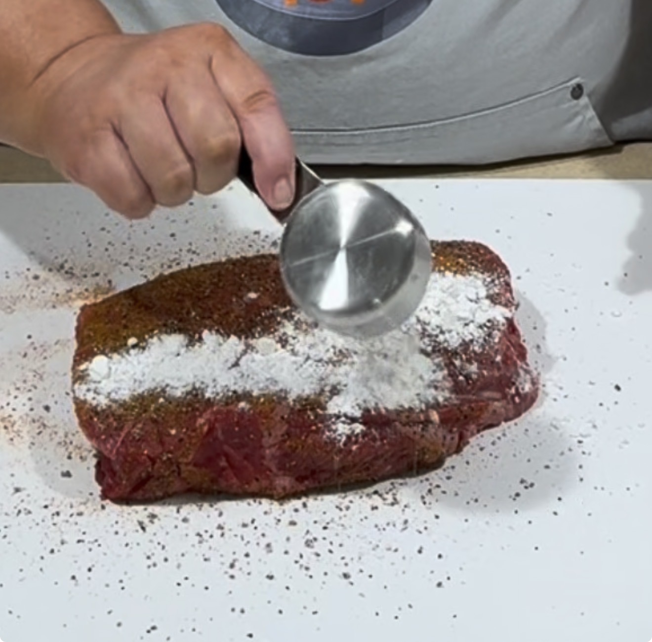 A person sprinkles flour from a metal measuring cup onto a seasoned raw beef roast resting on a white surface.