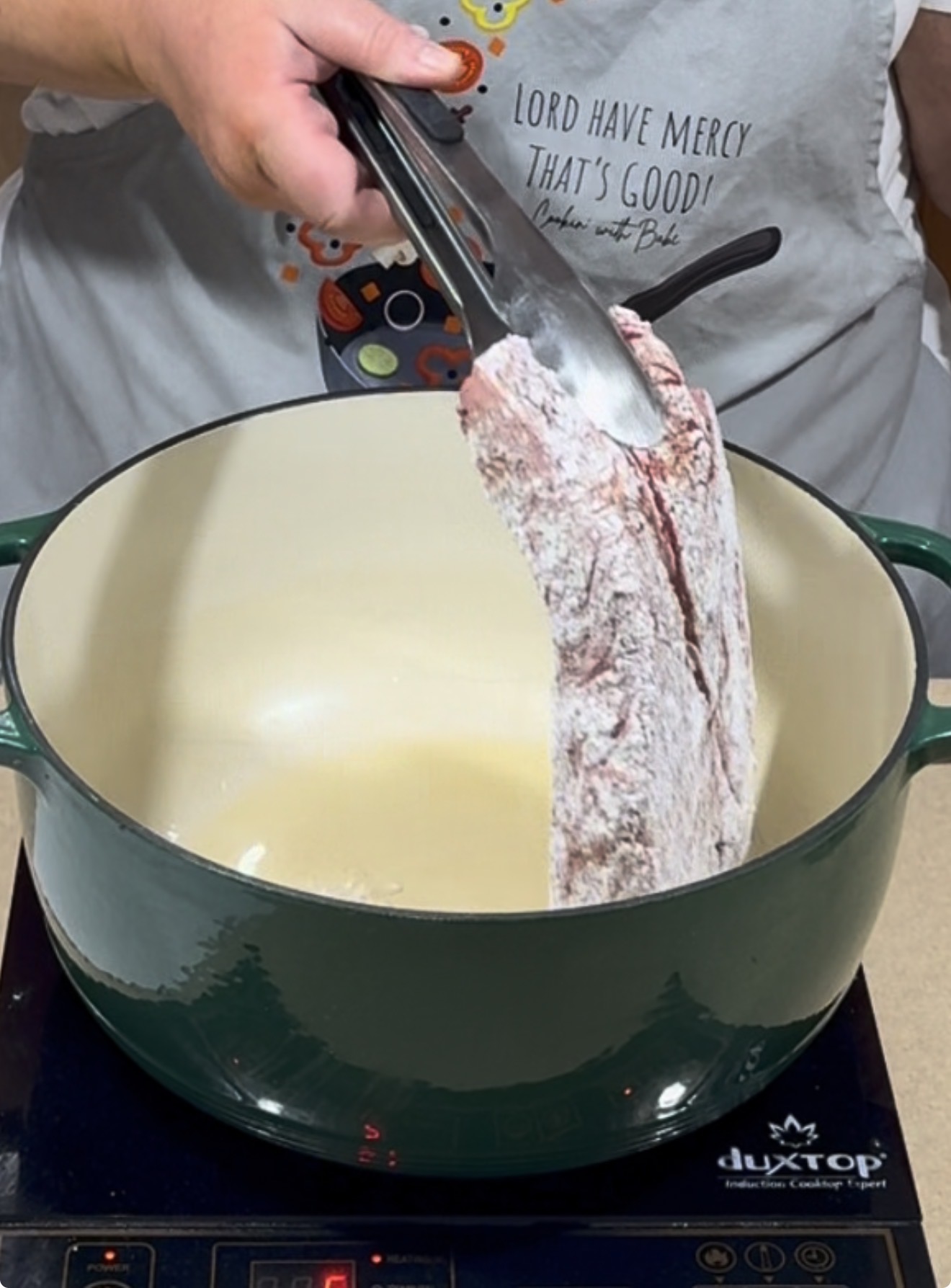 A person wearing an apron uses tongs to place a floured piece of beef into a large green Dutch oven on an induction cooktop.