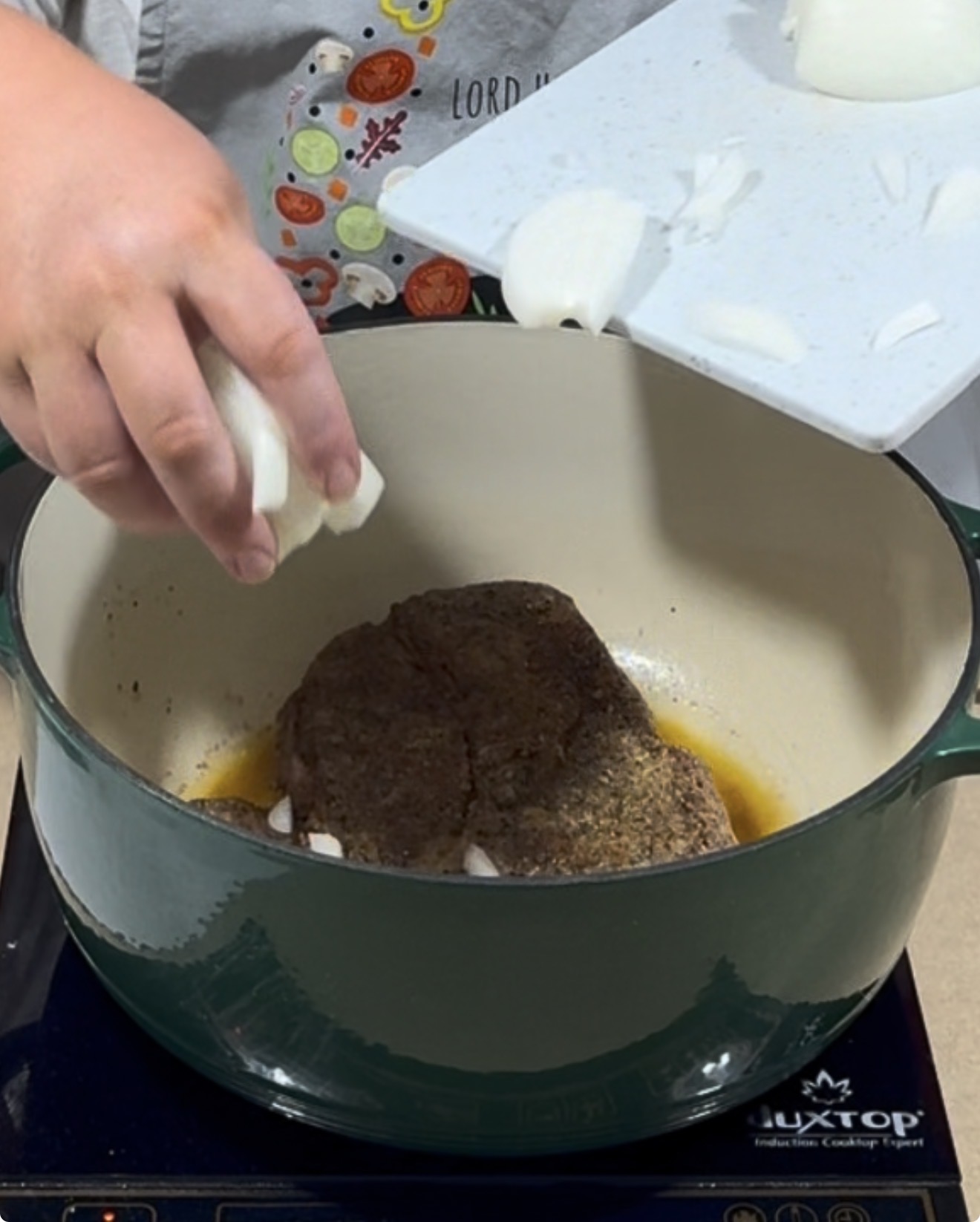 A person adds chopped onions from a white cutting board into a green Dutch oven with a large piece of seasoned meat browning inside.