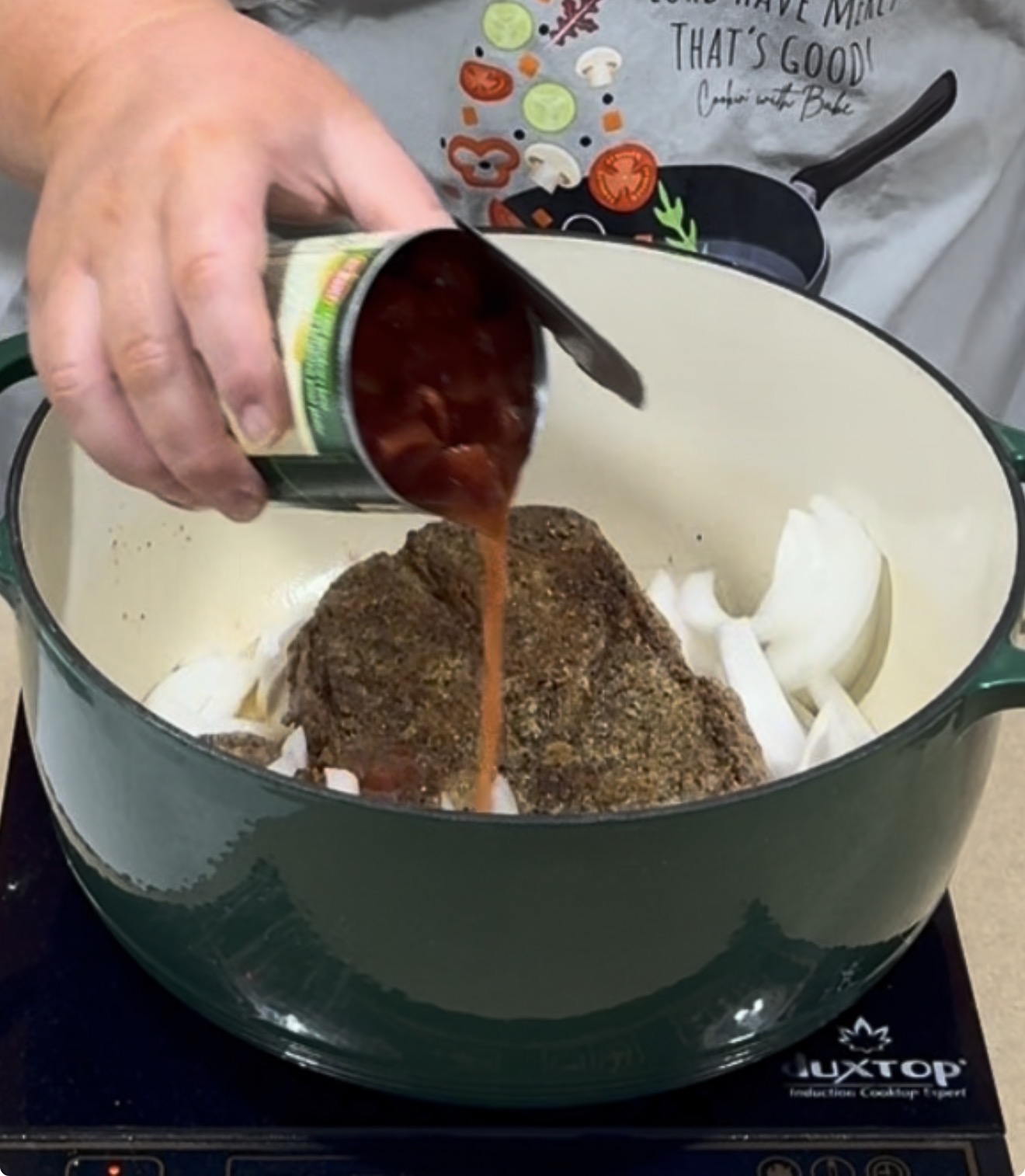 A hand pours canned diced tomatoes over a seasoned roast and sliced onions in a green Dutch oven on an induction cooktop.