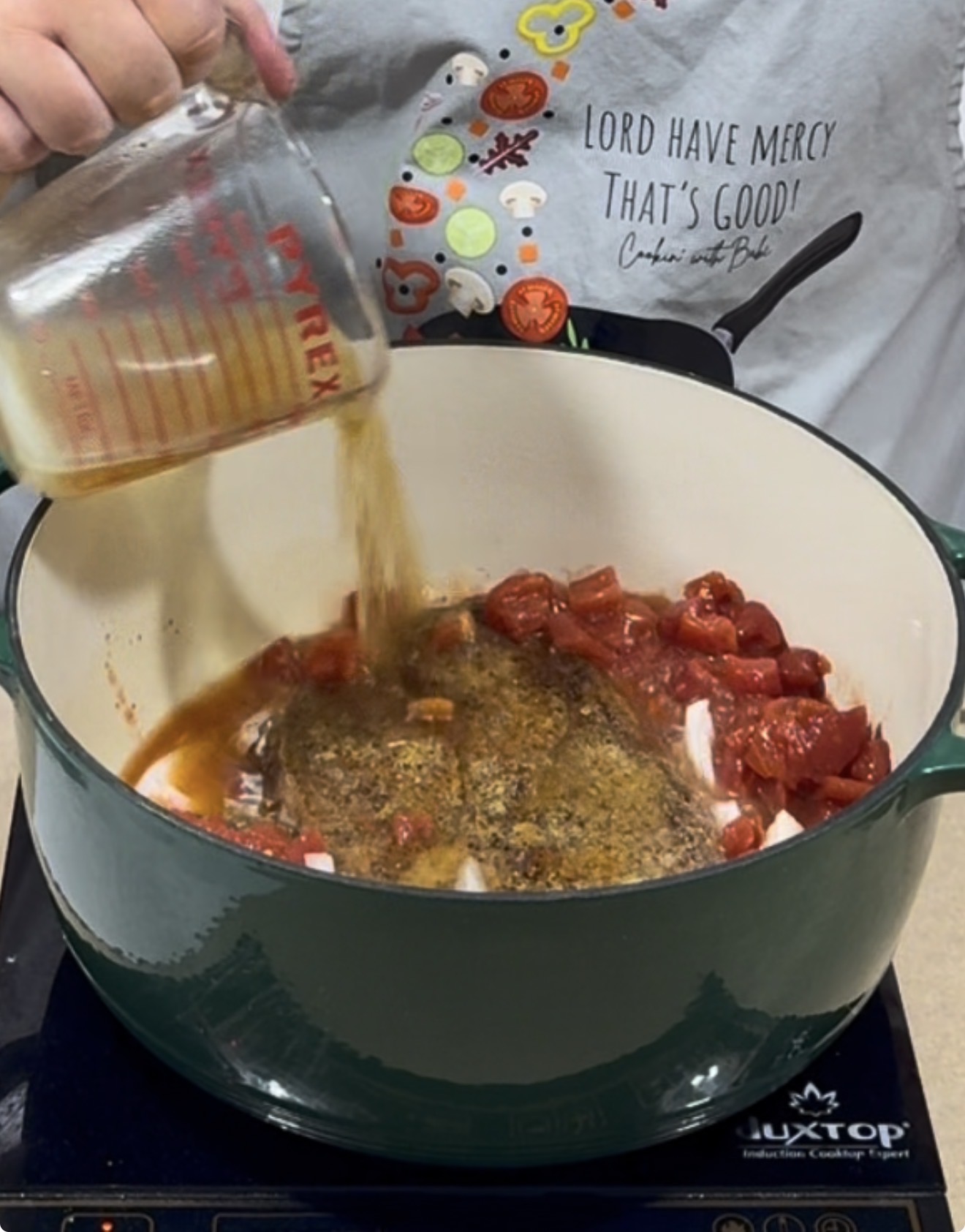 A person pours liquid from a measuring cup into a green pot with tomatoes and spices. The person wears a shirt with colorful icons and the phrase LORD HAVE MERCY THATS GOOD! Cookin with BAE..