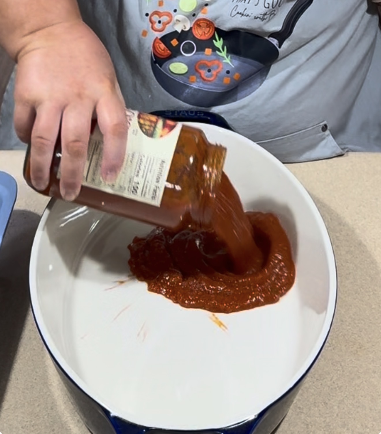 A person pours tomato sauce from a jar into a large white pot on a kitchen counter. Their apron has a colorful cooking-themed design.