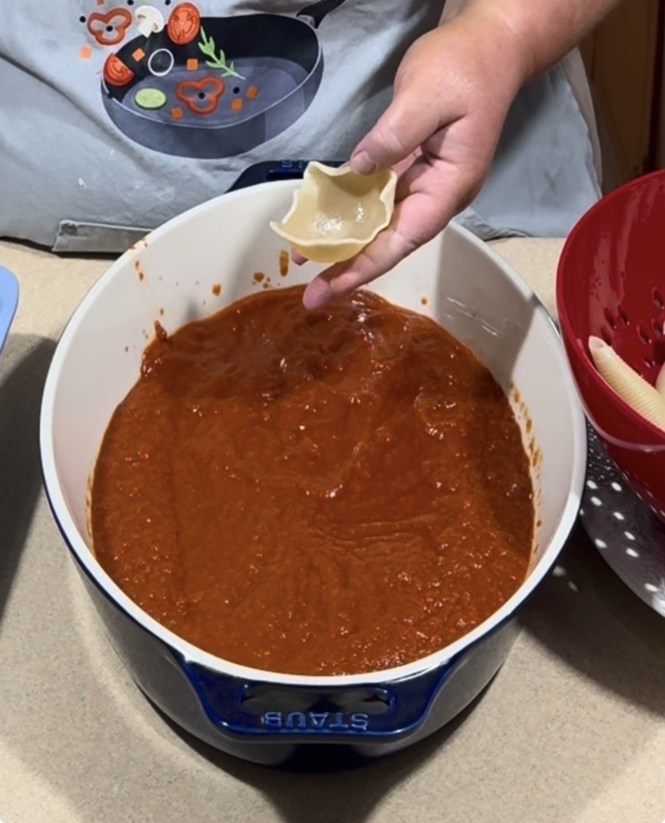 A person holds an uncooked stuffed pasta shell above a large blue pot filled with tomato sauce. Their apron has images of vegetables and a skillet on it.