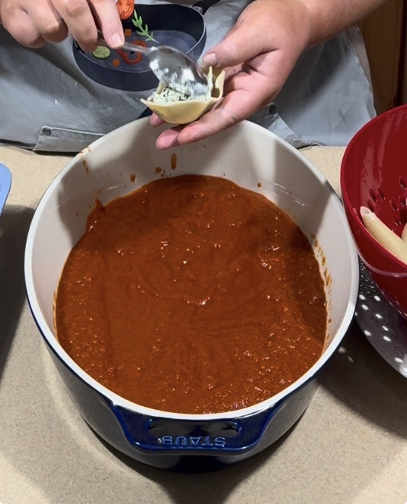 A person spoons ricotta filling into a pasta shell over a Dutch oven filled with red marinara sauce.