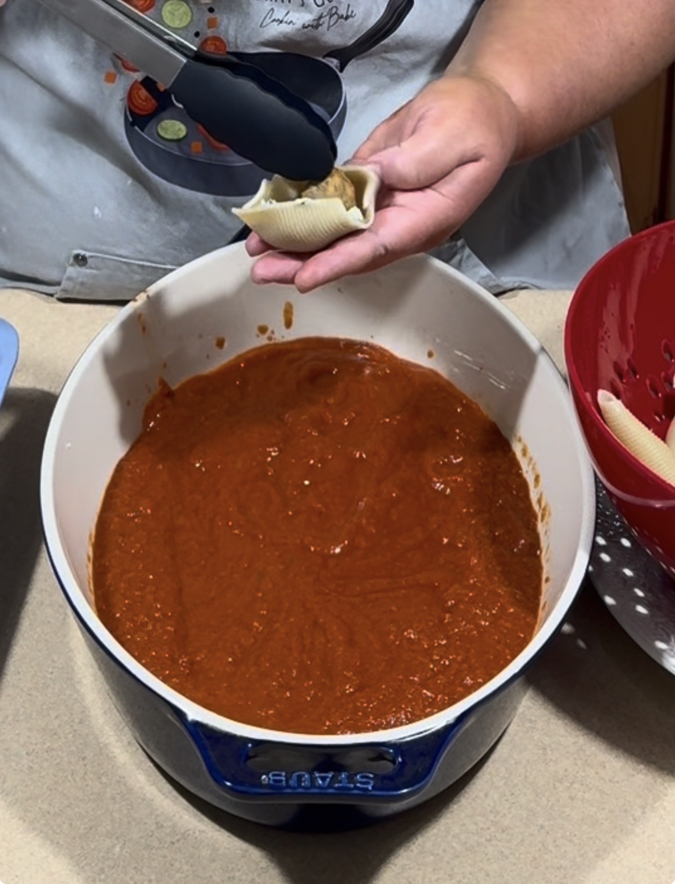 A person holds a pasta shell filled with cheese over a pot of red tomato sauce, preparing to add it to the dish. A red colander with more pasta shells is to the right.