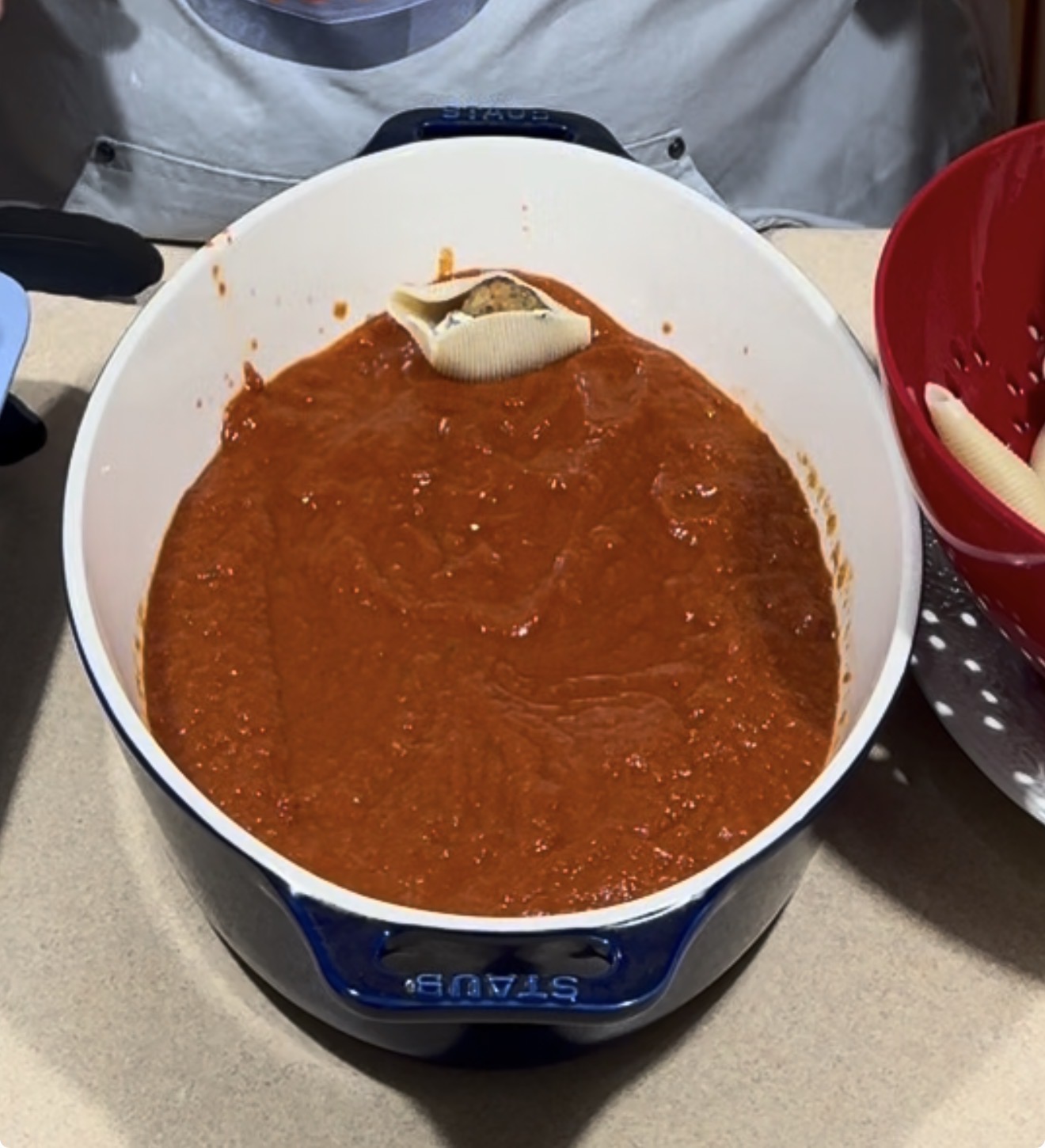A white Dutch oven filled with red tomato sauce contains a single stuffed pasta shell on top. A red colander with more pasta shells is partially visible to the right.