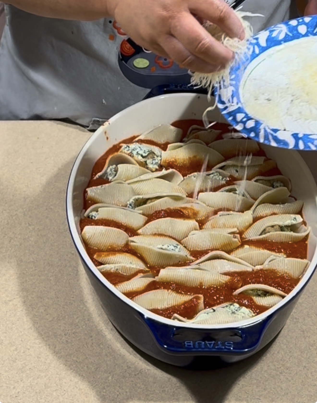 A person sprinkles shredded cheese onto stuffed pasta shells in a blue baking dish filled with tomato sauce, preparing the dish on a beige countertop.