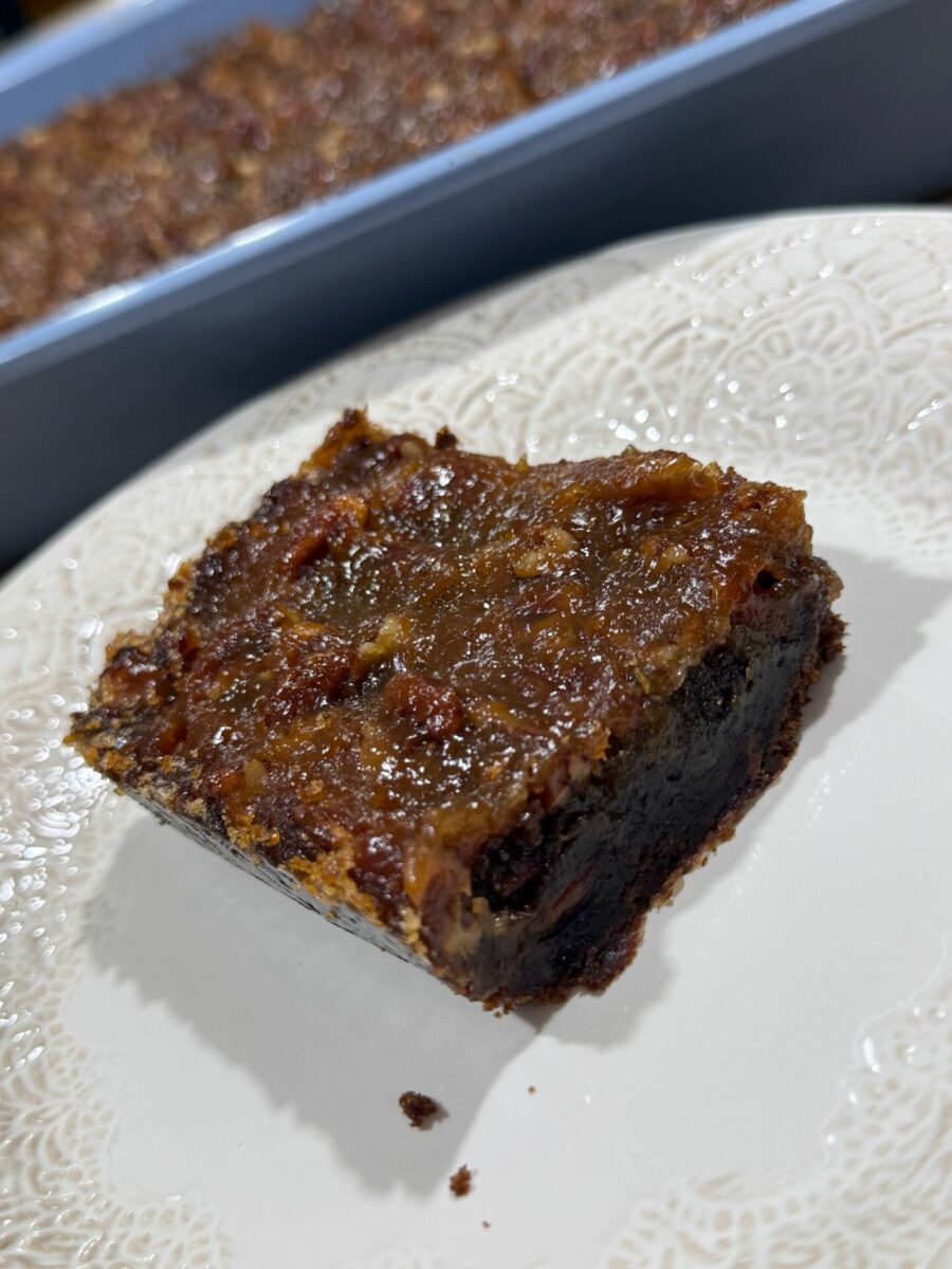 A close-up of a gooey dessert bar with a caramelized top layer, possibly pecan or date, on a white patterned plate. A tray of similar bars is visible in the background.