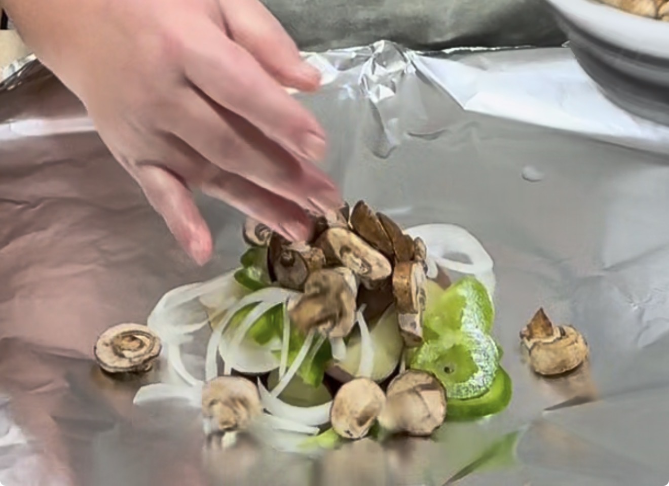 A hand places sliced mushrooms, onions, and green bell peppers on a sheet of aluminum foil, preparing vegetables for cooking.