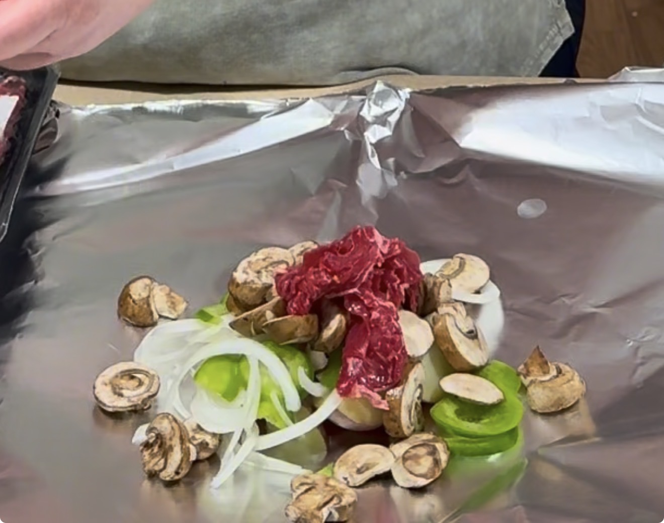 Sliced raw beef, mushrooms, onions, and green peppers are arranged on a piece of aluminum foil, ready for cooking. A persons hand is visible, preparing the ingredients.