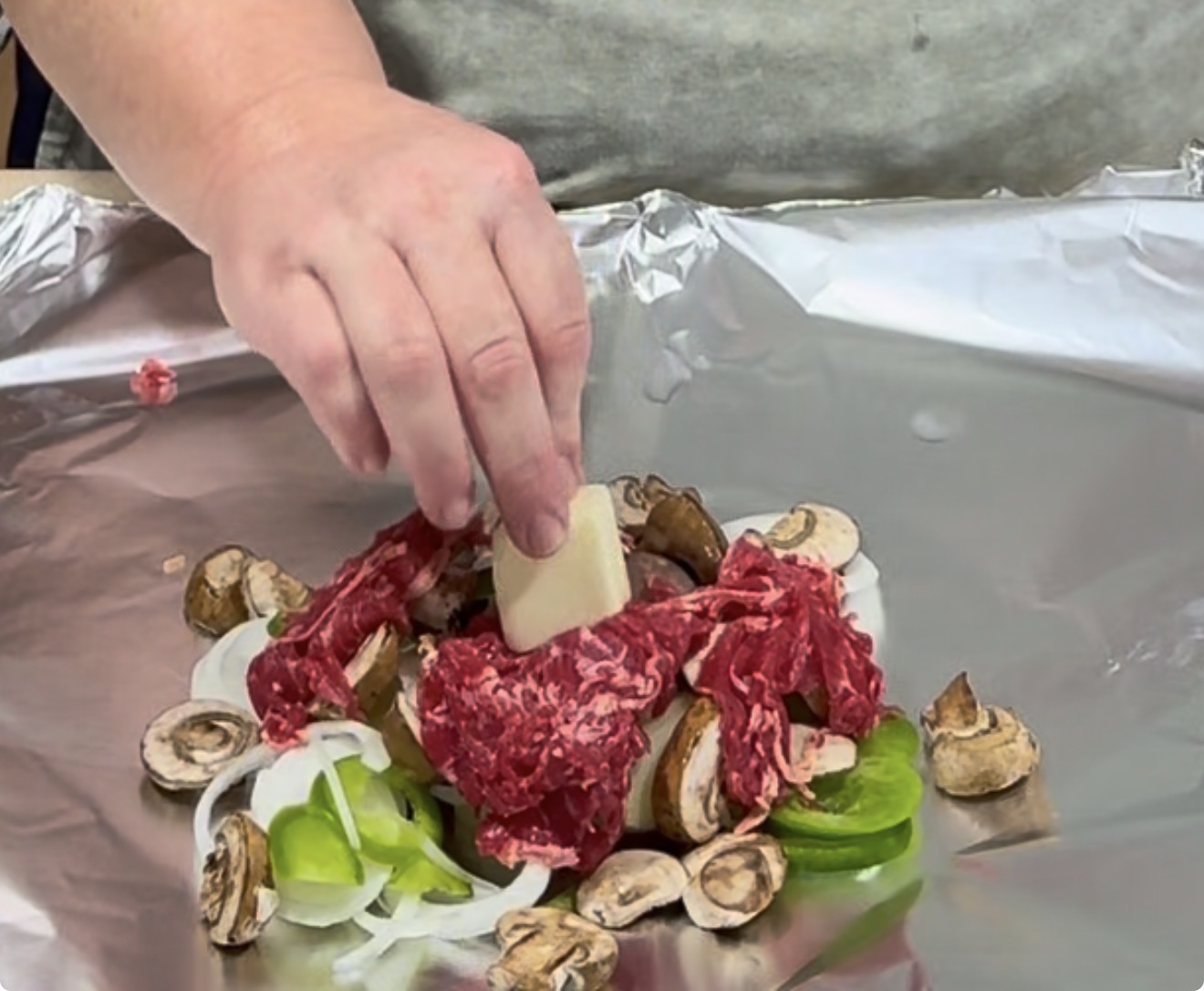 A hand places a piece of potato on top of sliced raw beef, mushrooms, onions, and green peppers arranged on a sheet of aluminum foil.