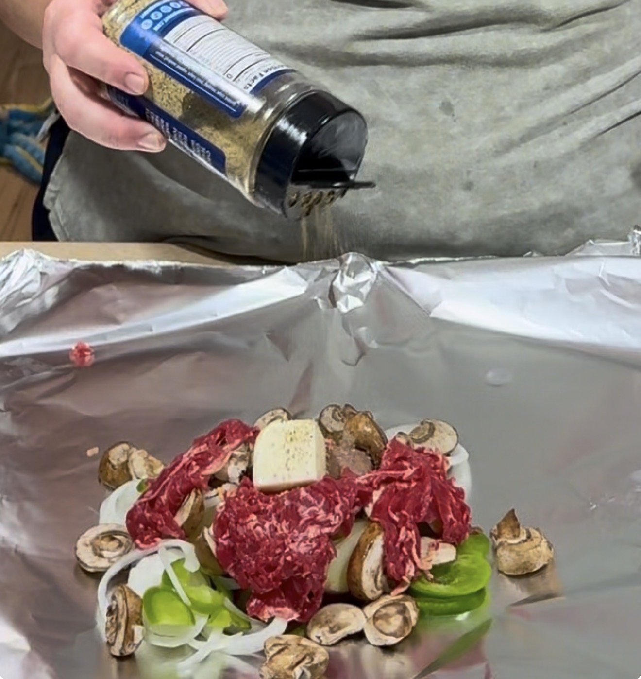 A person sprinkles seasoning onto sliced beef, mushrooms, onions, and peppers arranged on a sheet of aluminum foil.