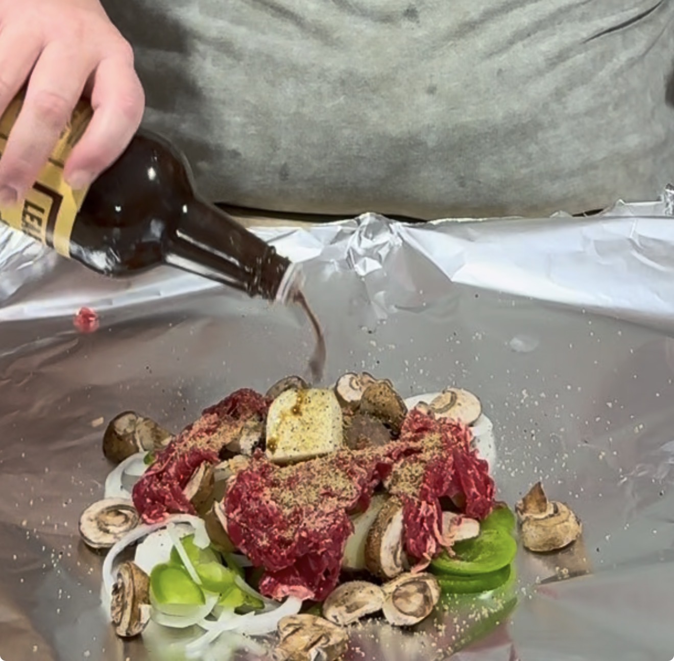 A person pours sauce from a bottle over raw beef, mushrooms, onions, and green bell peppers arranged on a sheet of aluminum foil.