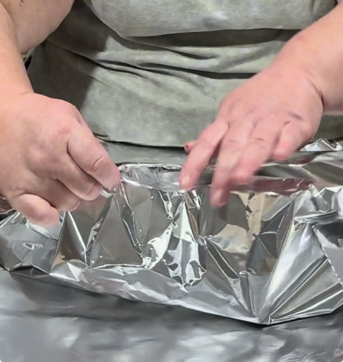 A person wraps or seals a large sheet of aluminum foil around an object on a table, using both hands.