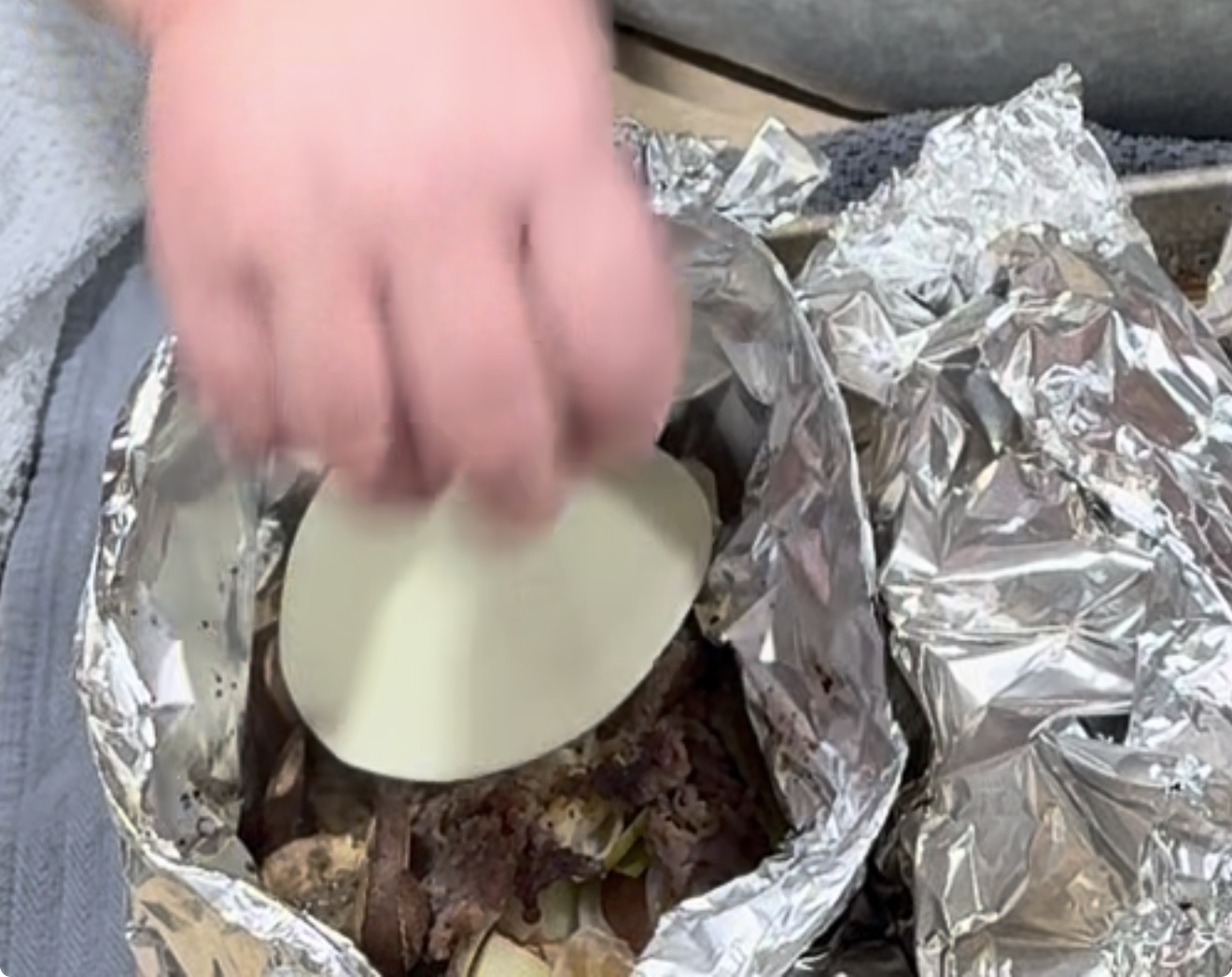 A hand holds a white disc over cooked meat and potatoes wrapped in crumpled aluminum foil. The food appears to be on a tray, and part of a blue towel is visible nearby.
