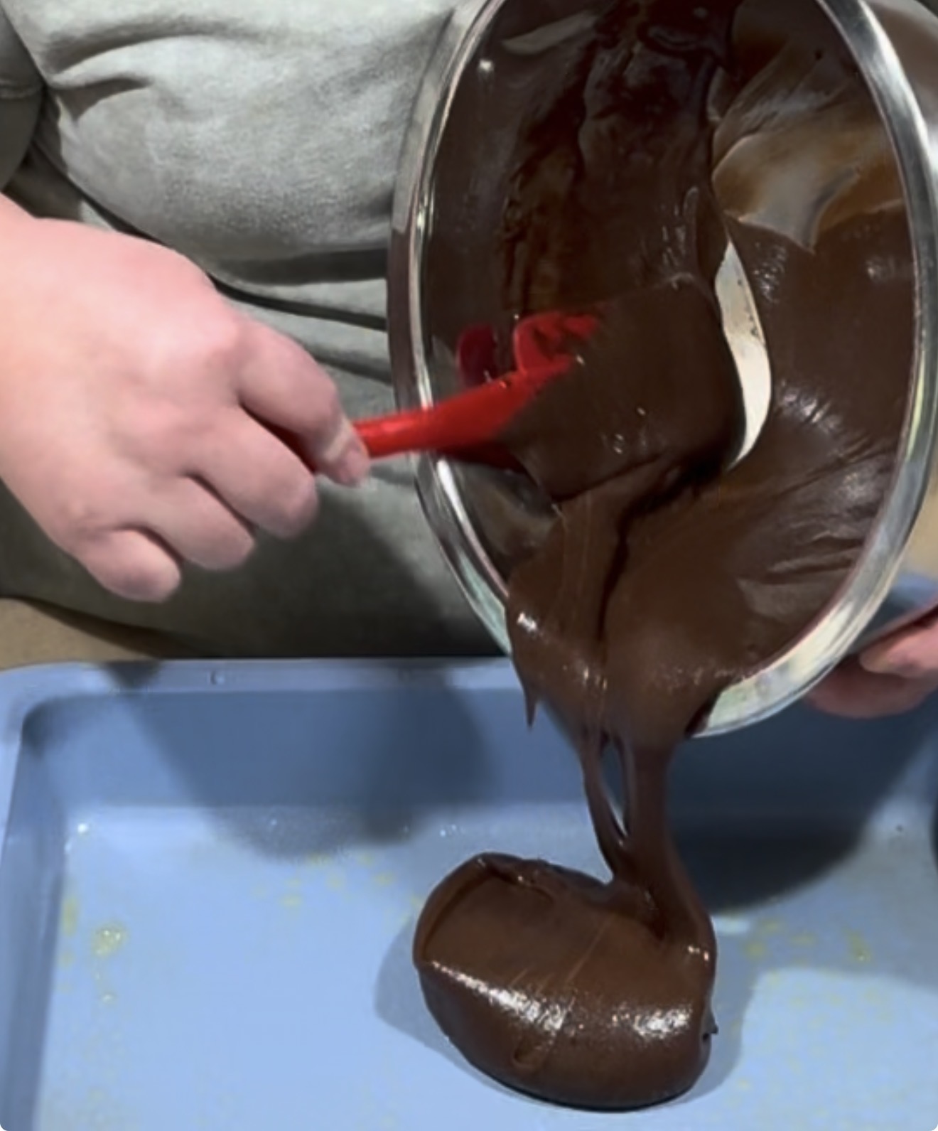 A person uses a red spatula to pour thick chocolate batter from a metal bowl into a blue baking pan.
