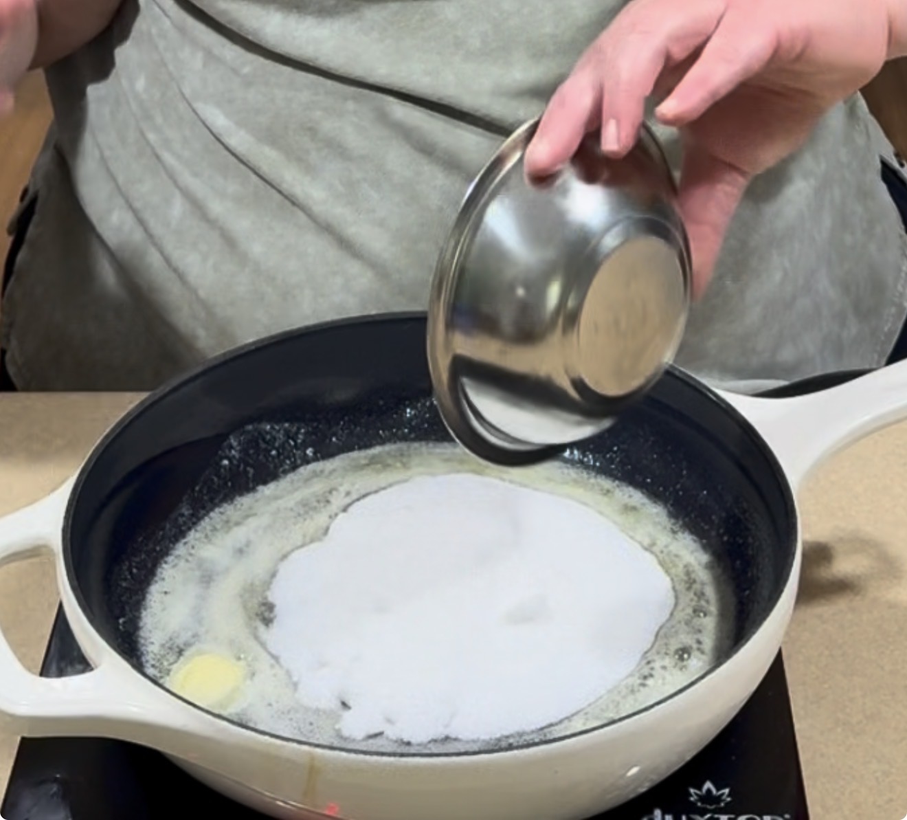 A person pours granulated sugar from a metal bowl into a white skillet with melted butter on a stovetop.