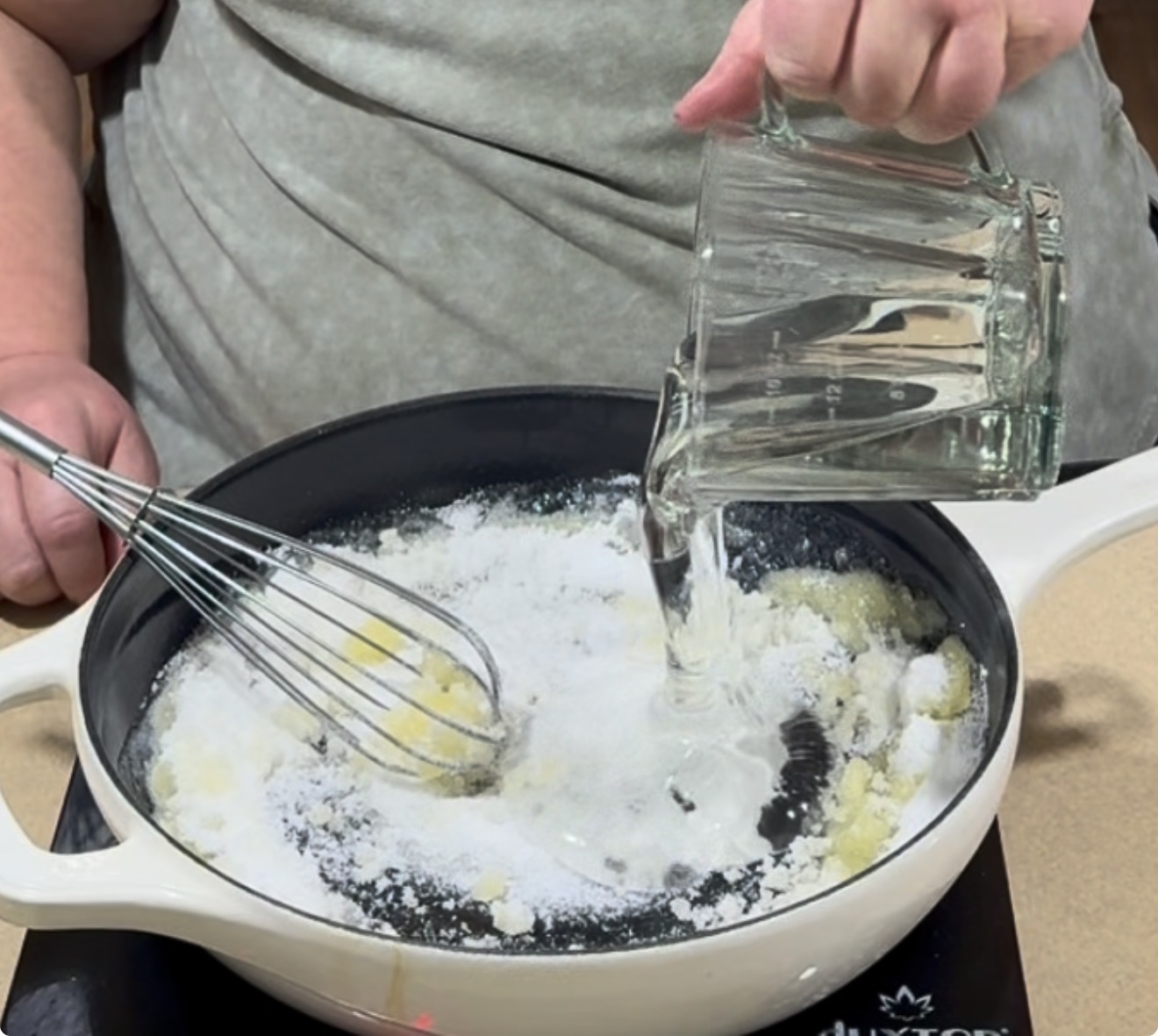 A person whisks white powder and butter in a white pan while pouring water from a glass pitcher onto the mixture.