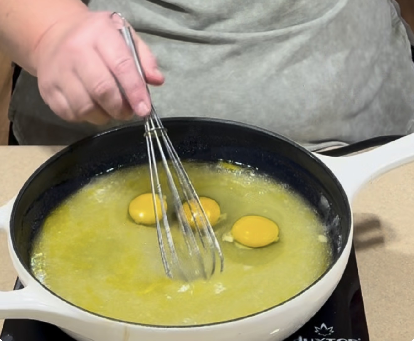 A person whisks three egg yolks into melted butter in a white pan on a stovetop, preparing to mix the ingredients.