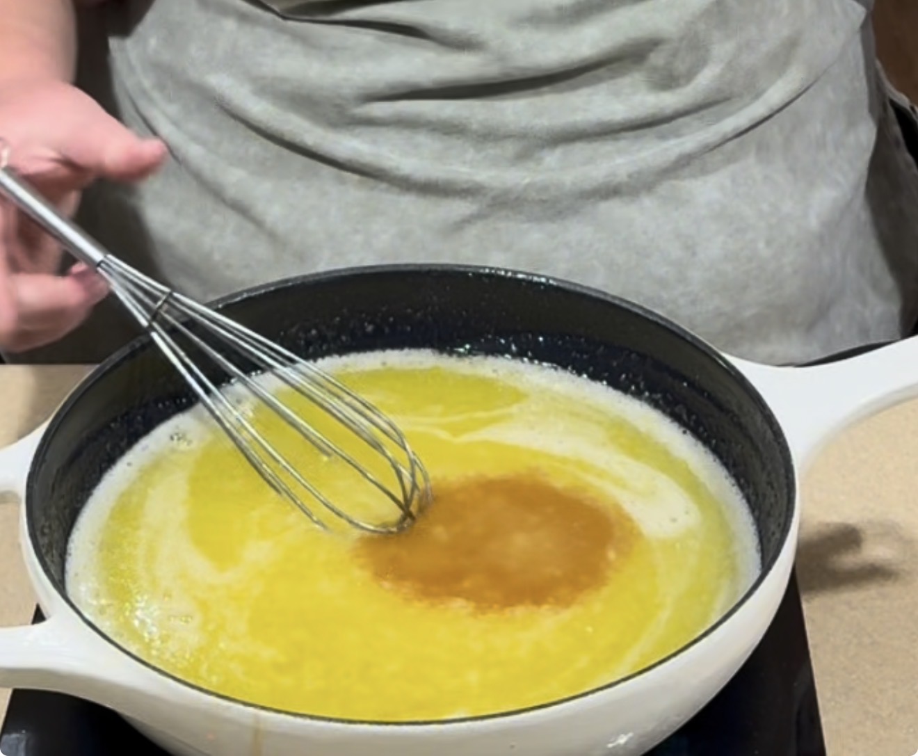 A person holds a whisk above a white pan filled with melted butter, which is being stirred on a stovetop.