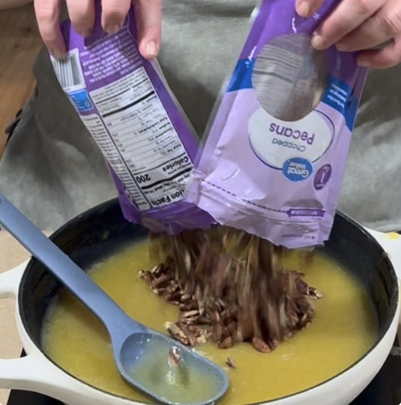 A person pours chopped pecans from two purple bags into a skillet filled with yellow liquid. A blue spoon rests in the pan.