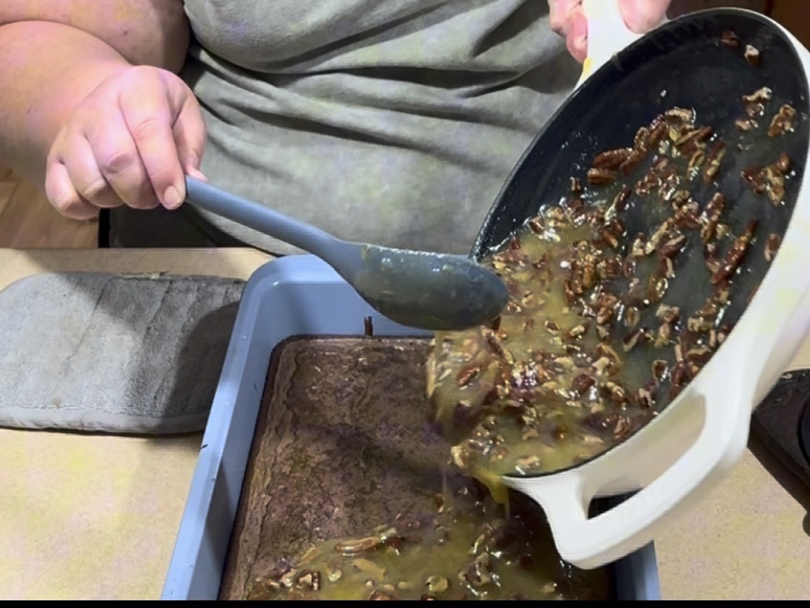 A person pours a nutty caramel mixture from a saucepan onto a baked brownie layer in a rectangular baking pan, using a gray spatula to help spread the topping.