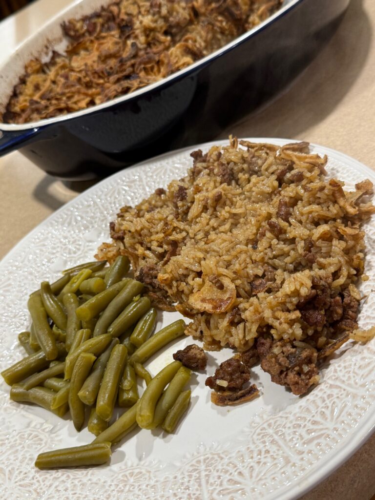A white plate with green beans and a serving of beef and rice casserole sits on a counter. In the background, more casserole is visible in a baking dish.
