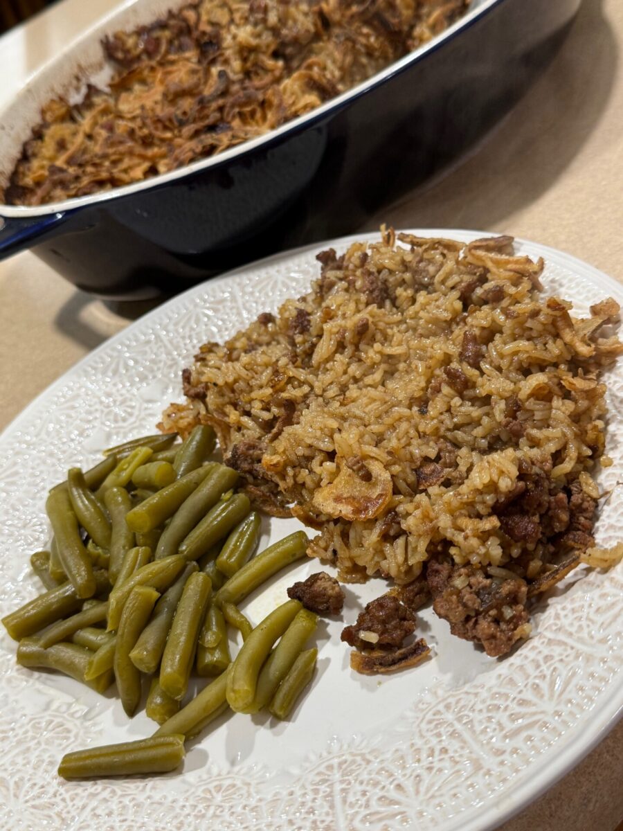 A white plate with green beans and a serving of beef and rice casserole sits on a counter. In the background, more casserole is visible in a baking dish.