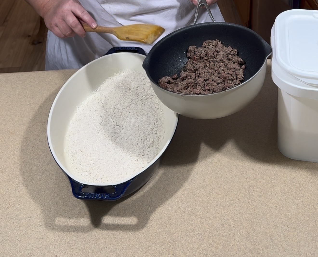 A person pours cooked ground beef from a frying pan into a white oval dish filled with uncooked rice, using a wooden spoon. A large container, likely holding rice, sits on the counter nearby.