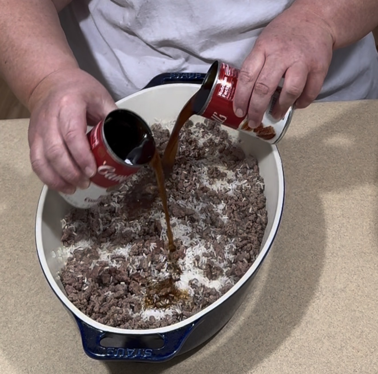 A person pours soup from two cans into a casserole dish filled with cooked ground beef and uncooked rice on a kitchen counter.