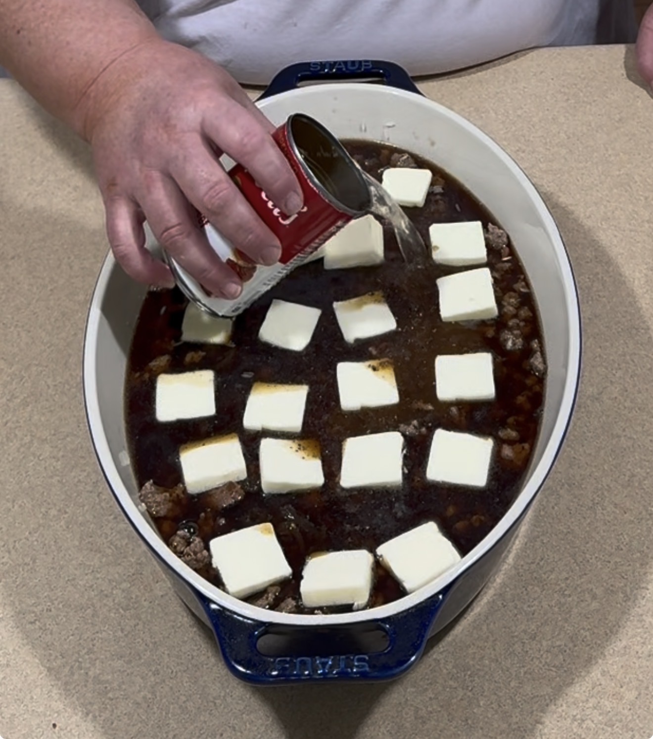 A person pours liquid from a can into a blue casserole dish filled with a dark mixture, ground meat, and evenly spaced butter squares on top, set on a beige countertop.