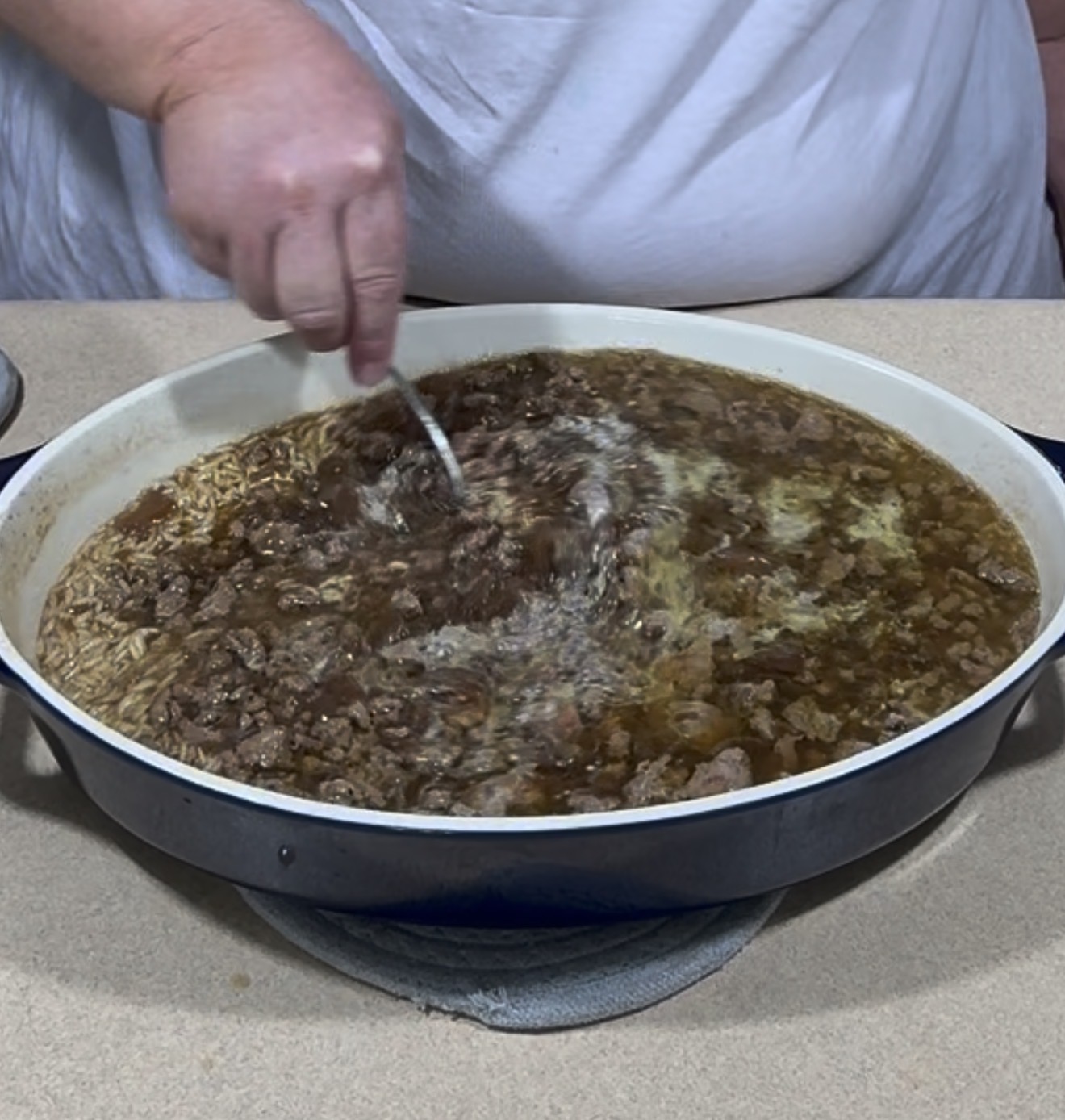 A person wearing a white shirt stirs a large pan of beef and rice in broth, which is placed on a beige countertop.