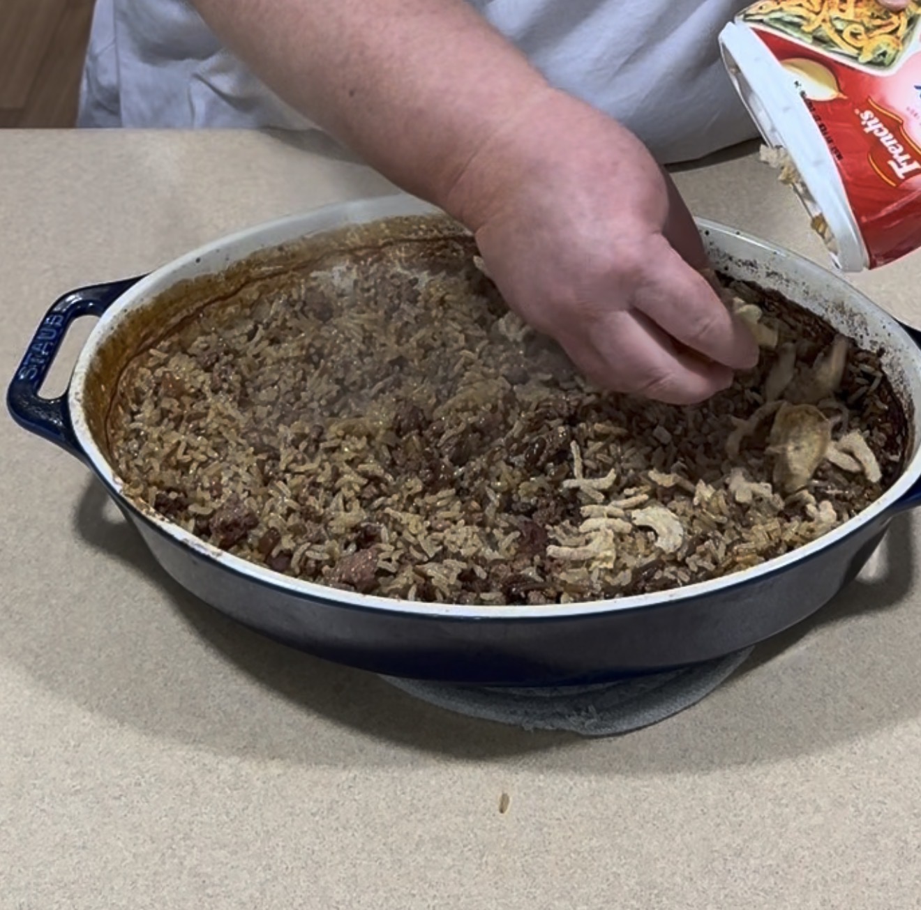 A person sprinkles fried onions from a can onto a baked rice and meat casserole in a blue oval dish on a countertop.