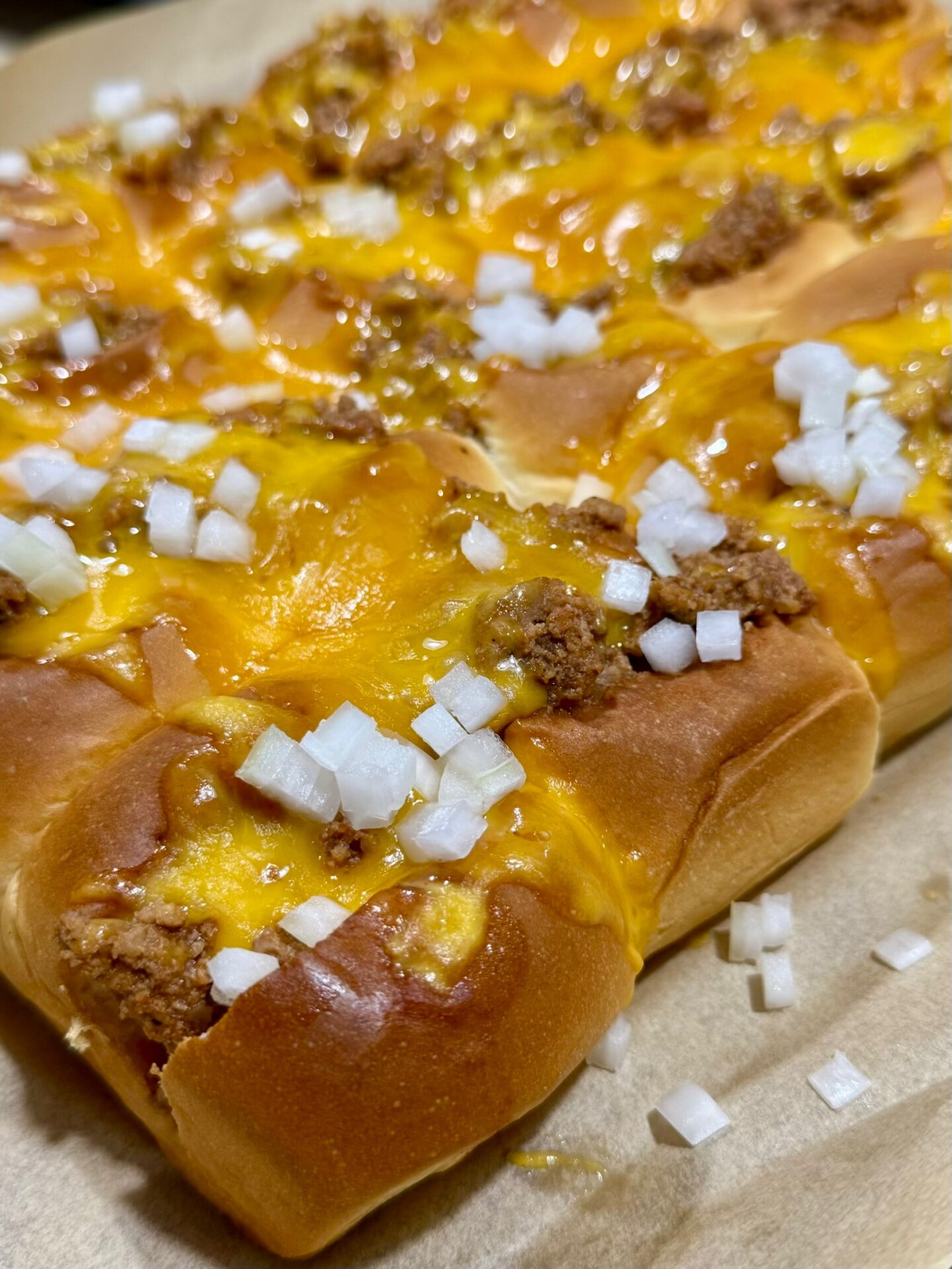 Close-up of baked hot dog buns topped with melted cheddar cheese, chopped onions, and crumbled ground meat, all on parchment paper.