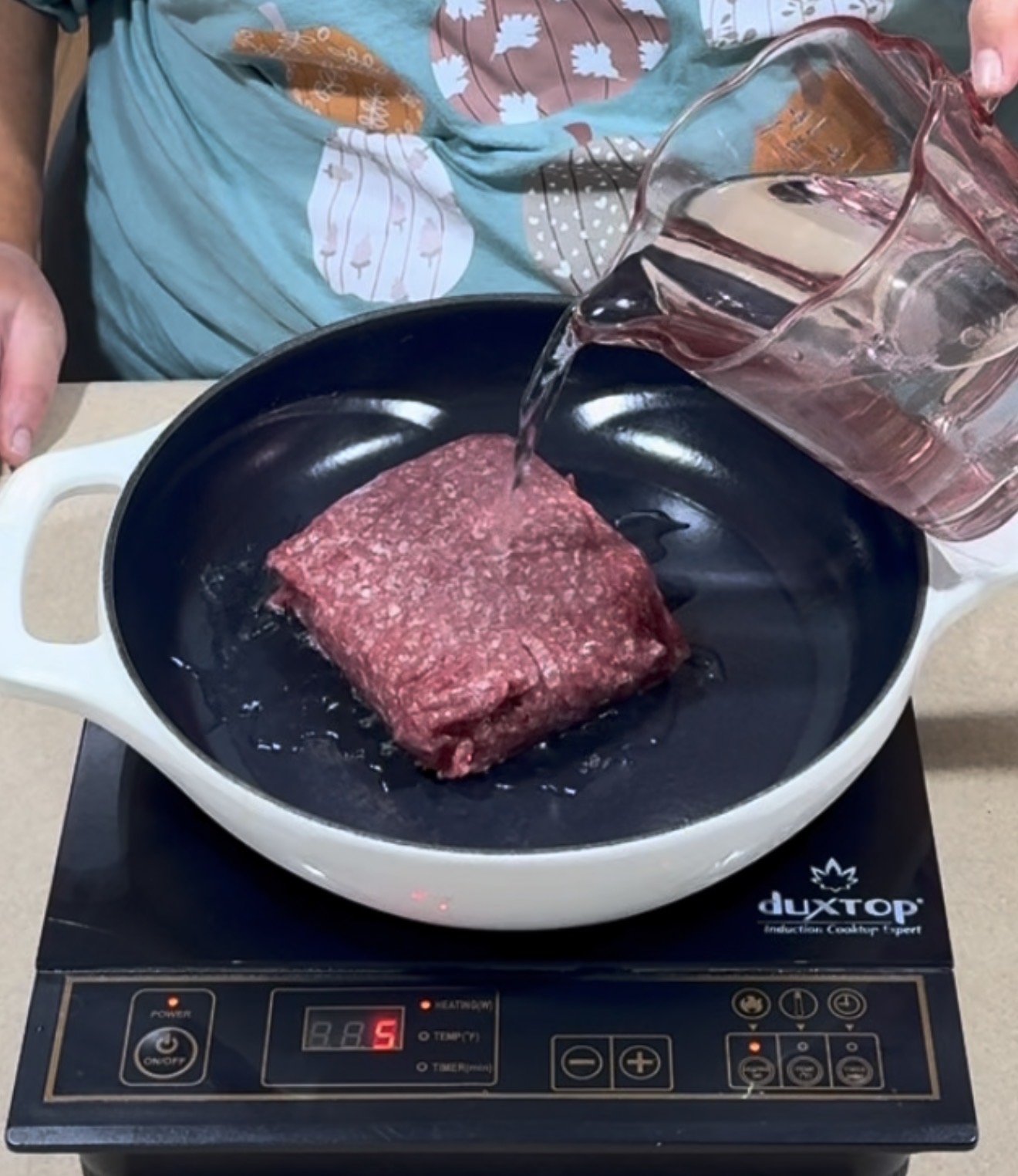 A person pours water from a measuring cup into a pan with raw ground beef on an electric cooktop.