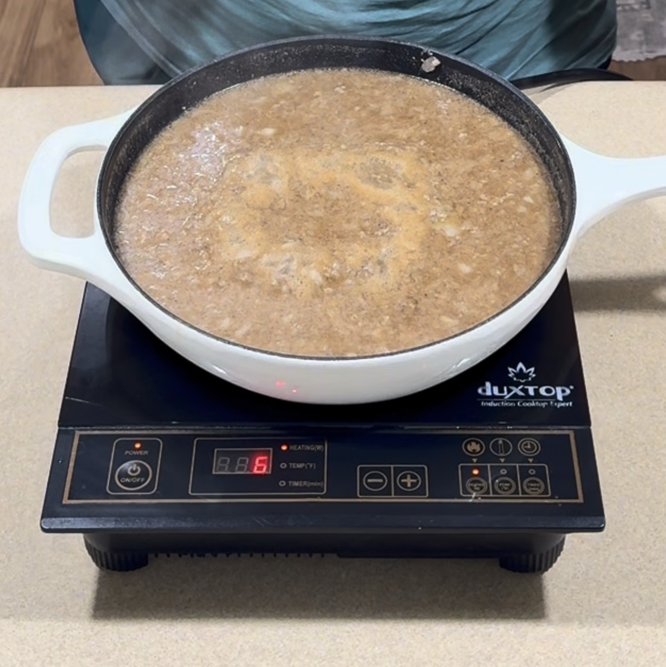 A white pot on a Duxtop induction cooktop is filled with simmering brown soup or sauce. The display shows level 6, and the cooktop is placed on a beige countertop.