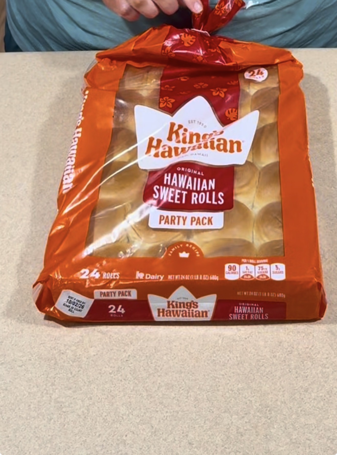 A person opens a 24-pack bag of Kings Hawaiian Original Sweet Rolls labeled Party Pack on a beige countertop.