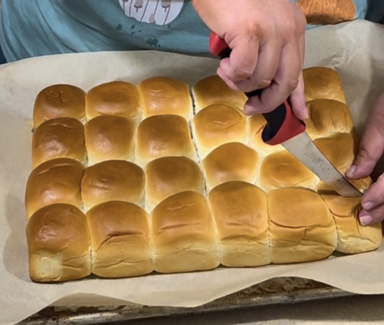 A person uses a knife to cut a sheet of golden brown dinner rolls on a parchment-lined baking tray.